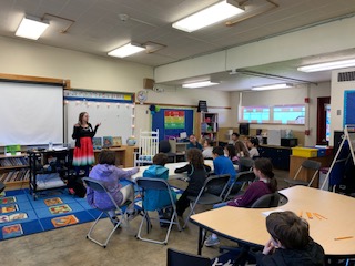 Cindy teaching in front of a classroom. She is wearing a watermelon dress and there are several students in the room.